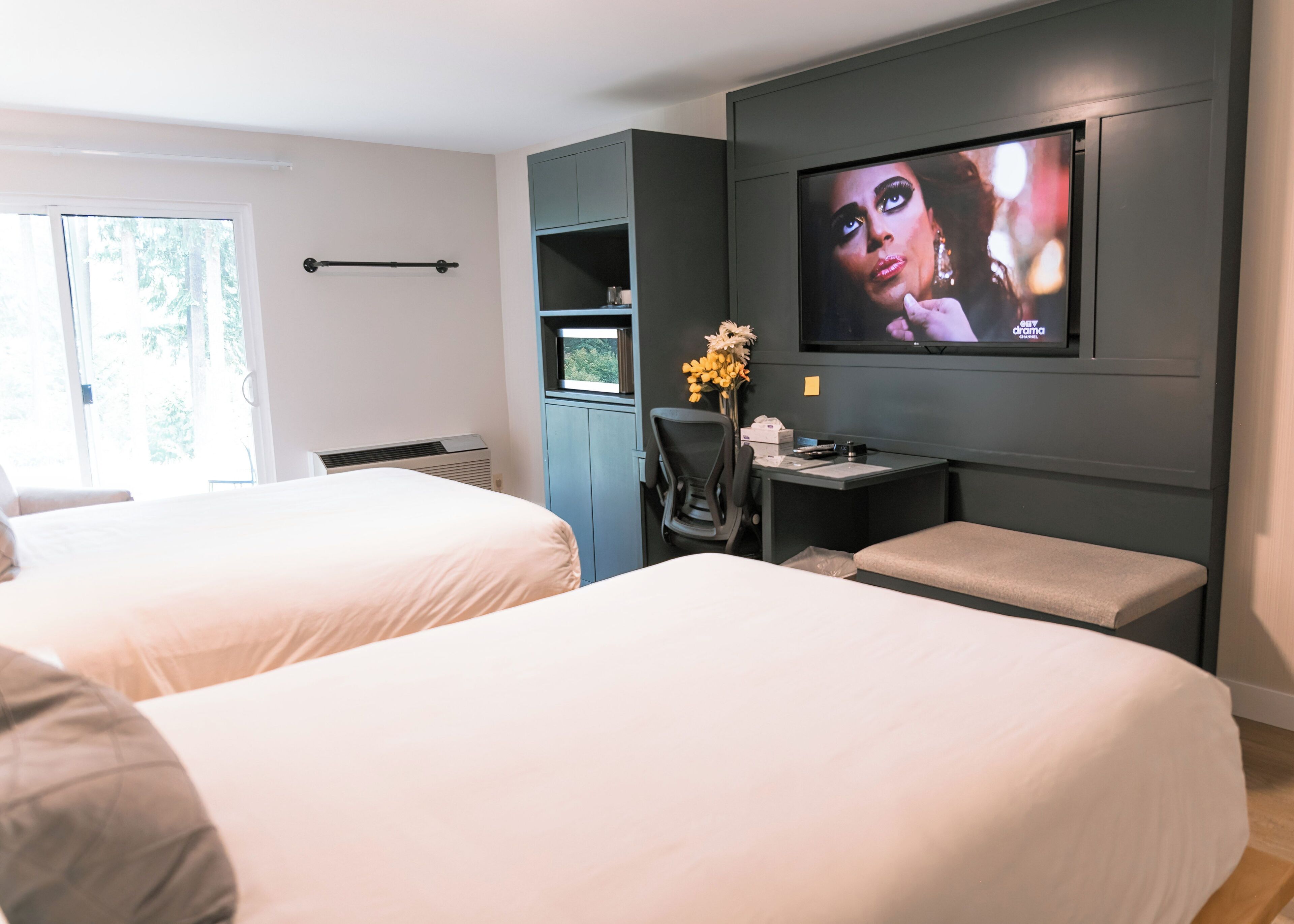 A hotel room with two beds, a desk with a TV, and a window with a view of trees.