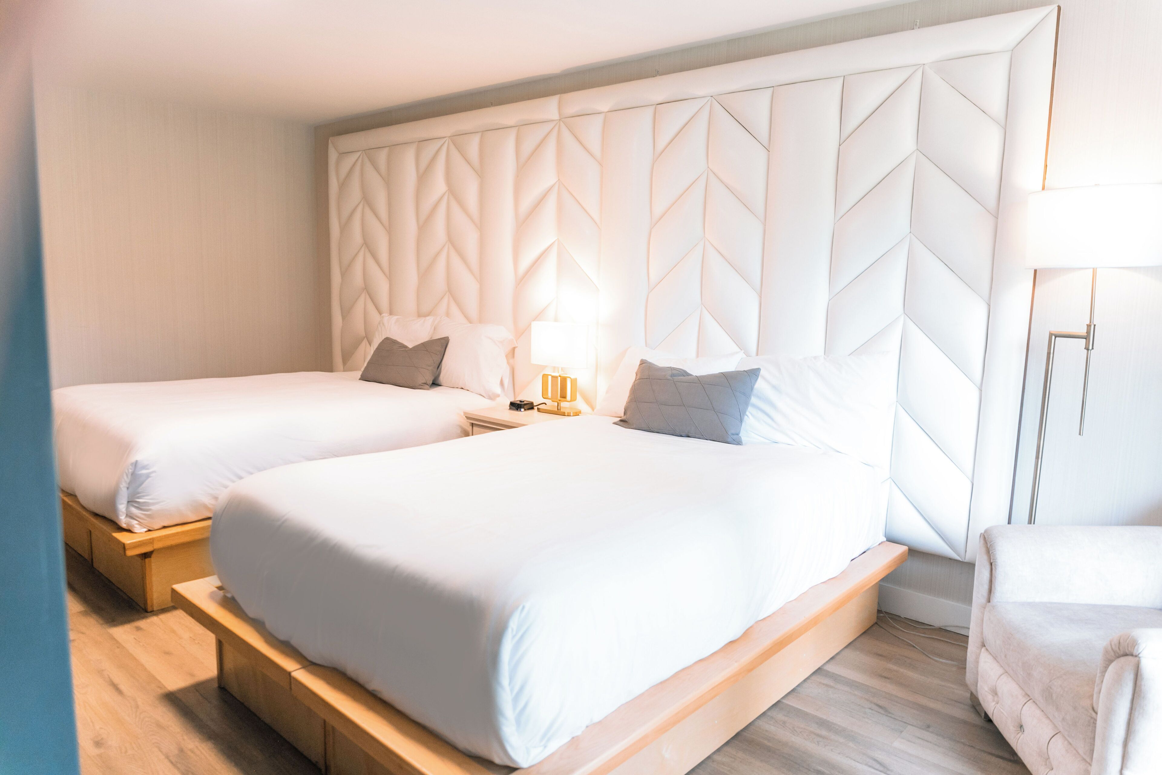 A hotel room with two beds, a large padded headboard, a bedside table with a lamp, and a chair.