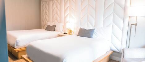 A hotel room with two beds, a large padded headboard, a bedside table with a lamp, and a chair.