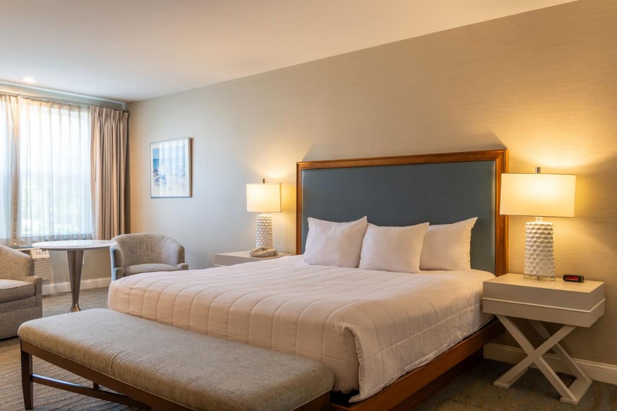 A hotel room with a large bed, two bedside tables, a bench, a chair, and a window with curtains.