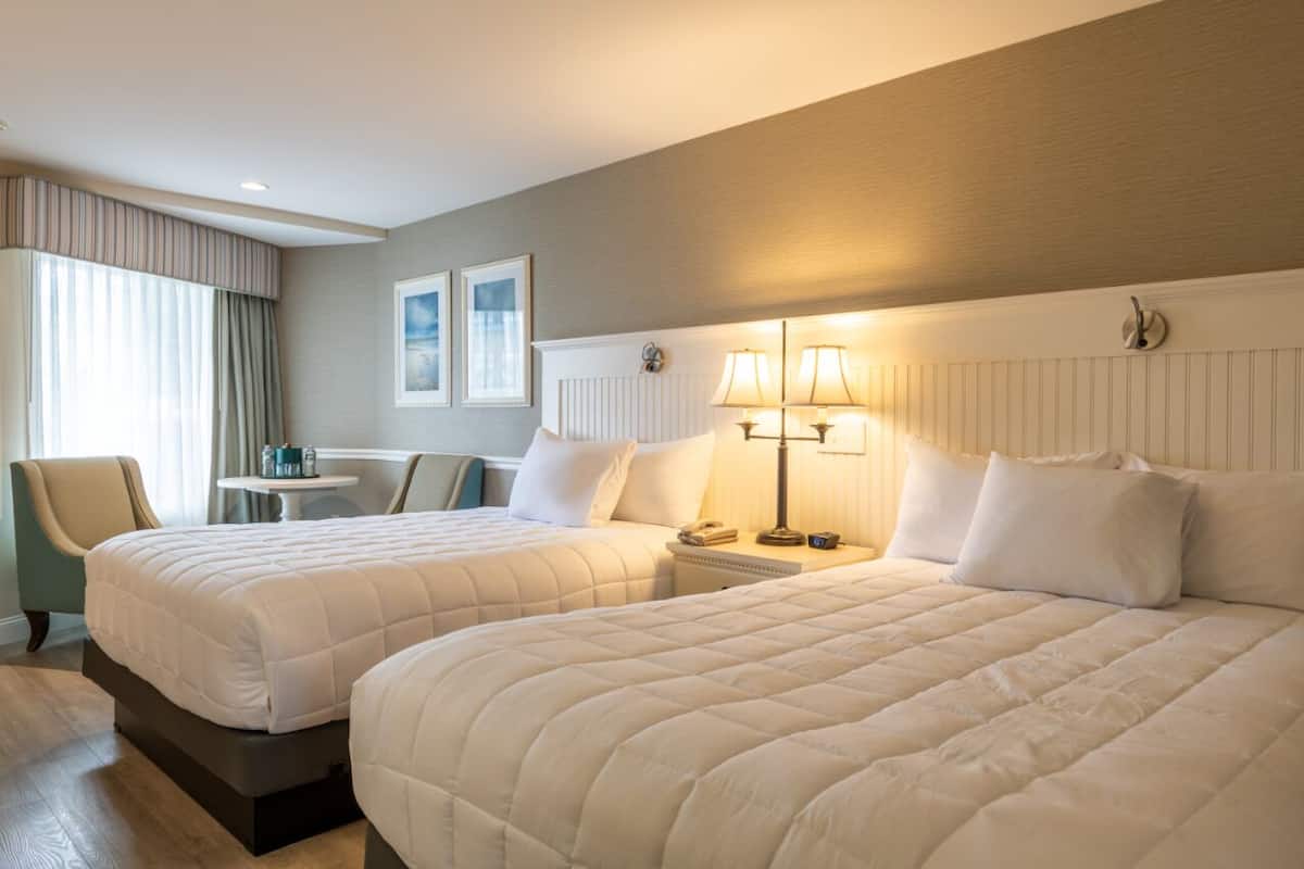 A hotel room with two beds, a small table, a chair, and a lamp.