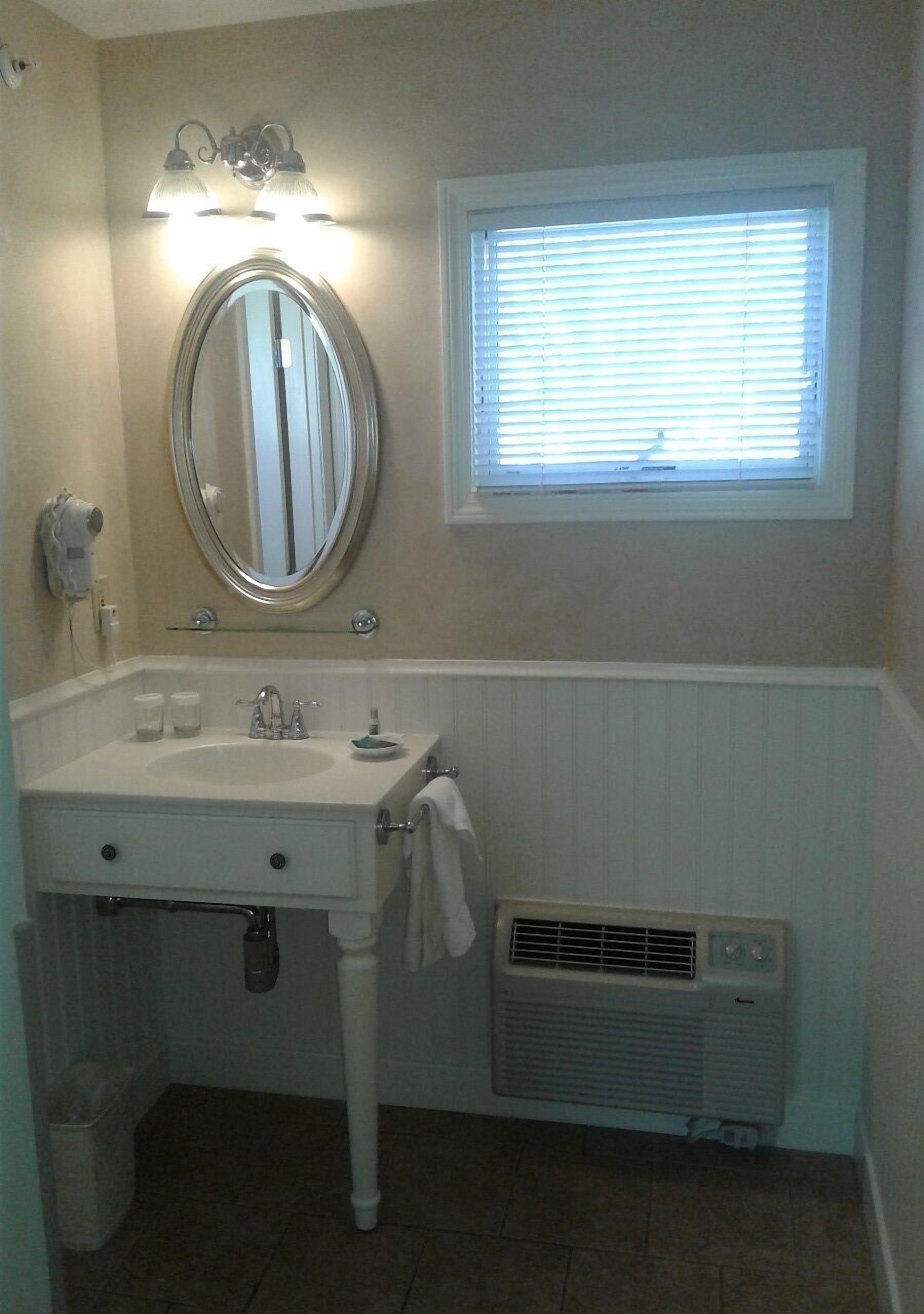 A bathroom with a white vanity, a round mirror, a window with blinds, and a wall-mounted heater.