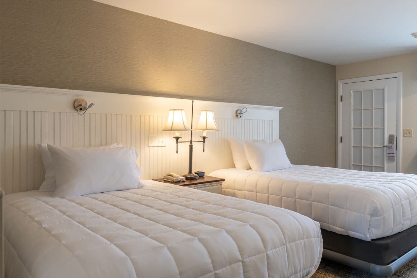 Two beds with white bedding, a nightstand with a lamp, and a door with a window.