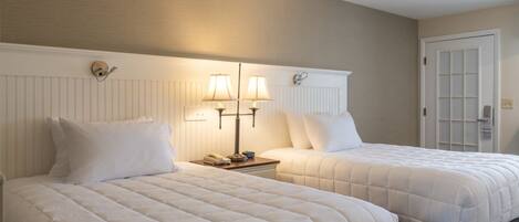 Two beds with white bedding, a nightstand with a lamp, and a door with a window.