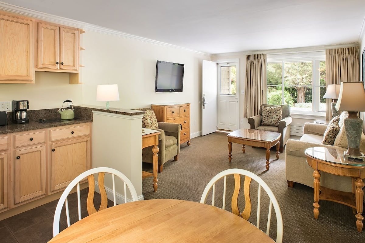 A compact hotel room with a kitchenette, a dining area, a living room with a sofa and armchairs, and a television mounted on the wall.