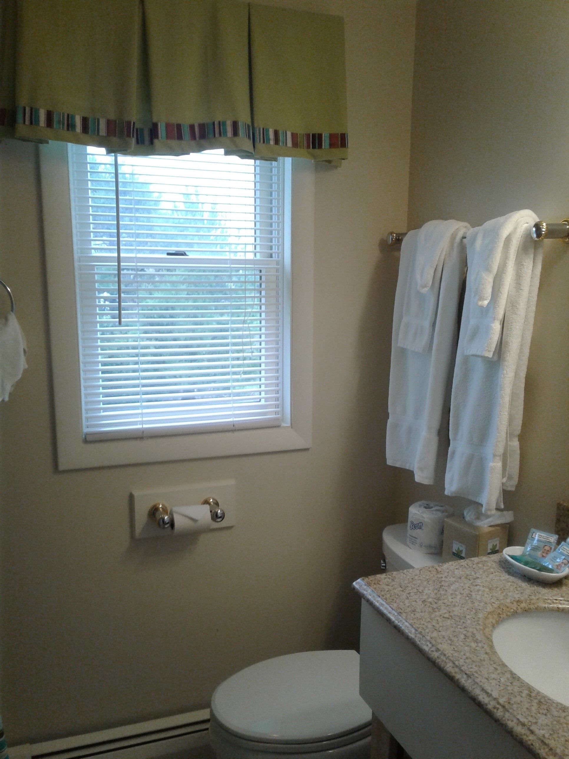 Une salle de bain équipée de toilettes, d’un lavabo et d’une fenêtre avec des stores. Des serviettes sont suspendues sur un porte-serviettes et un rouleau de papier toilette est fixé au mur.