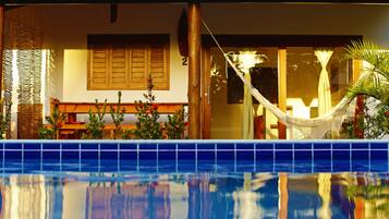 Vista junto a la piscina con una hamaca, contraventanas de madera y una piscina con baldosas.