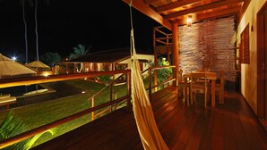 Una terraza de madera con una hamaca, una mesa y sillas, con vista a un césped cuidado y un edificio con un diseño de pared singular.