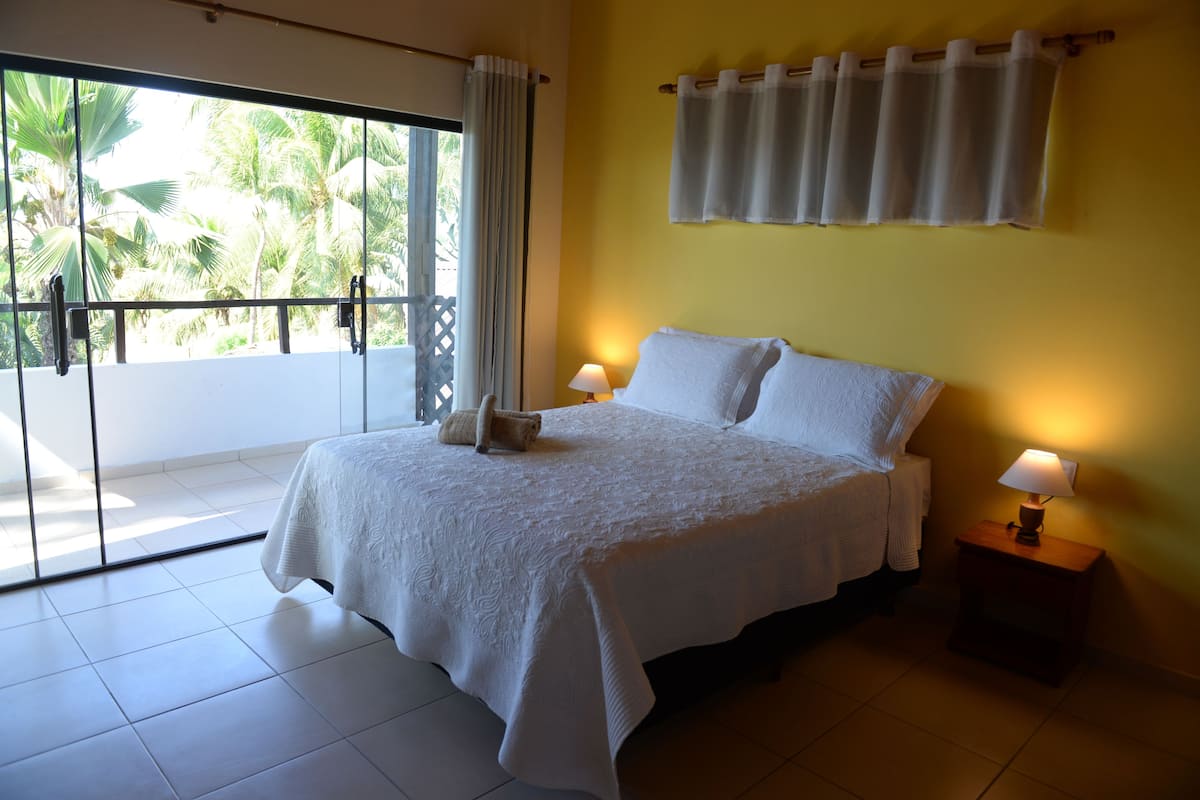 A bedroom with a bed, two bedside lamps, a window with curtains, and a view of palm trees.