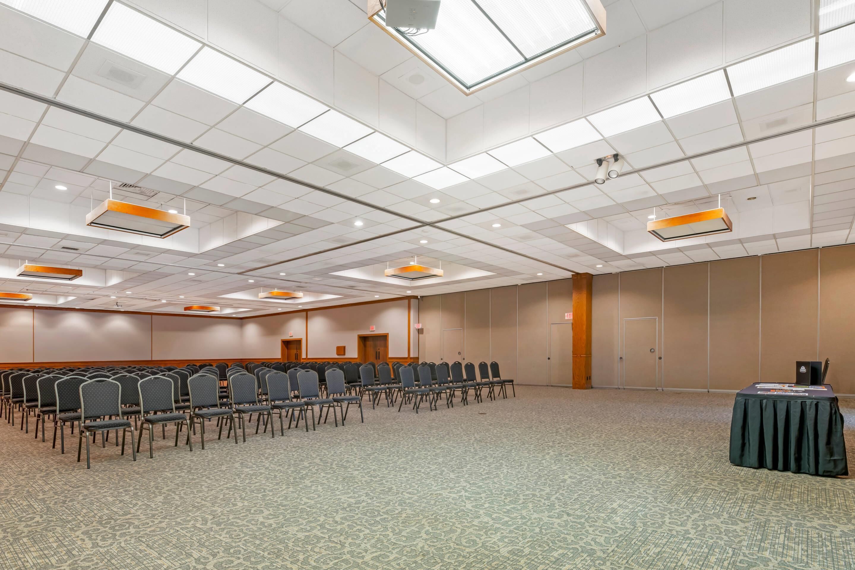 Una sala conferenze con file di sedie, un tavolo ricoperto da una tovaglia nera e un soffitto con luci a incasso e finestroni.