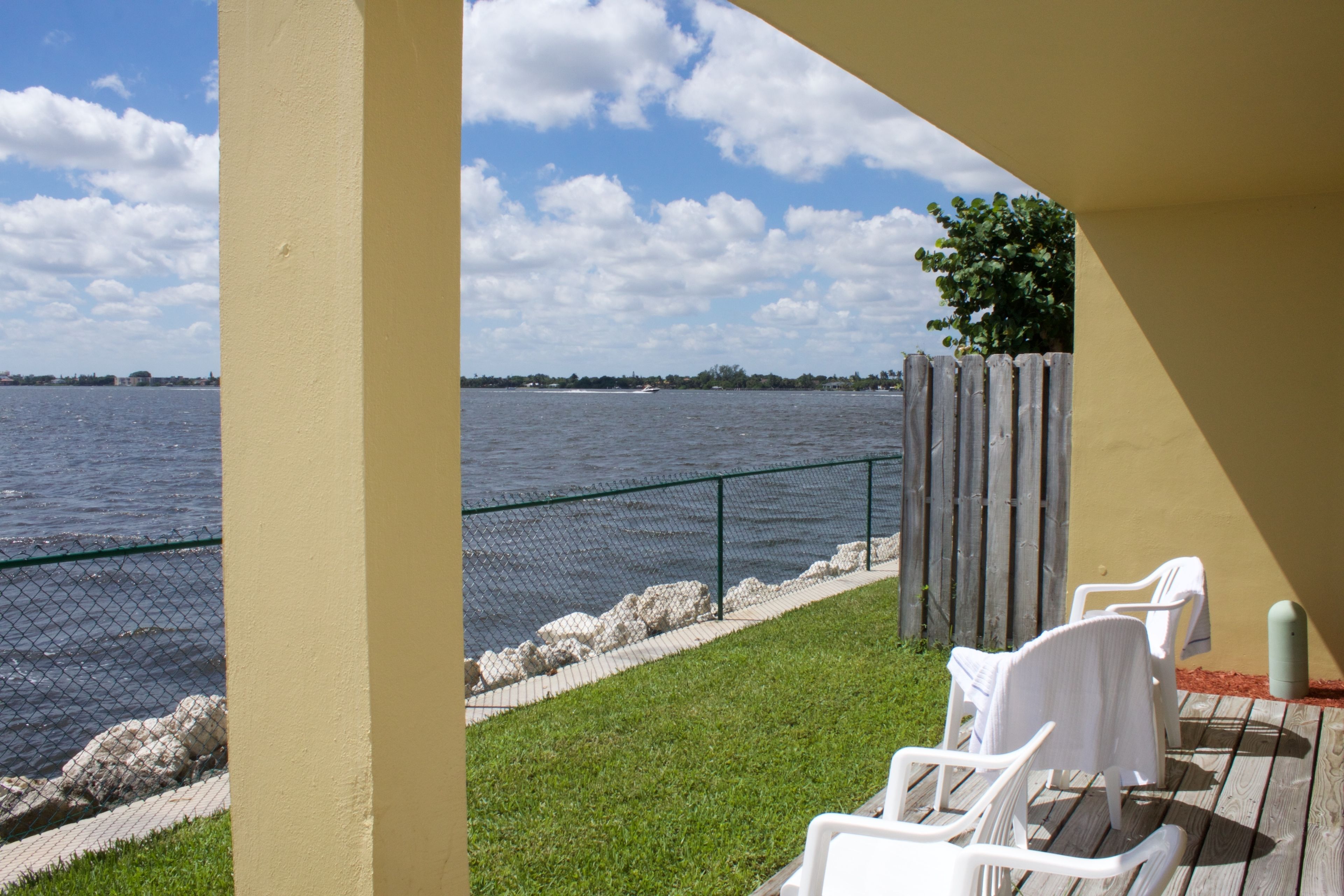 A patio with a view of the water, a fence, and two white chairs.