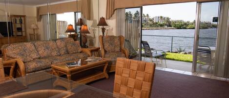 A living room with a sofa, armchairs, a glass coffee table, and a view of a body of water through large sliding doors.