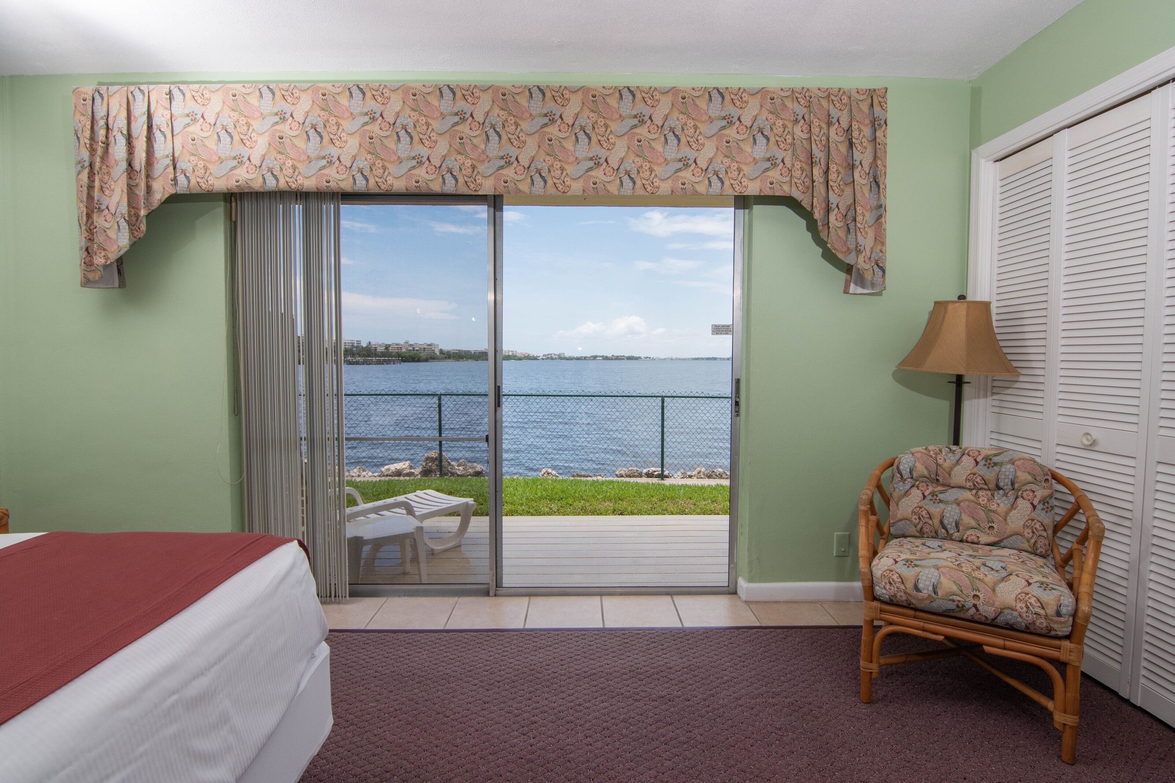 A room with a bed, a wicker chair, a view of the sea, and a balcony with a table and chairs.
