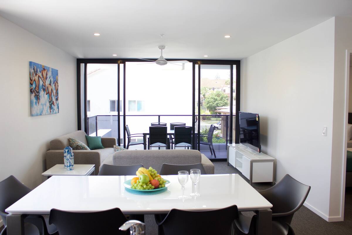 A modern living room with a dining area, a sofa, a TV, and a balcony with outdoor seating.