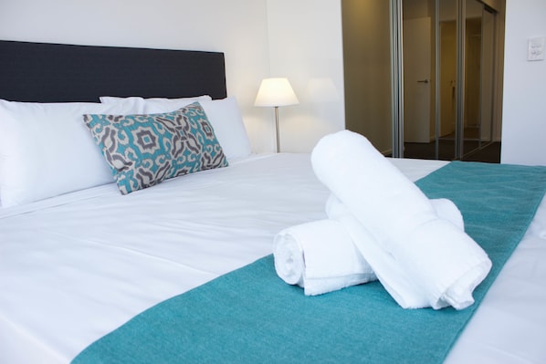 A neatly made bed with white linens, a teal throw blanket, and a rolled white towel.