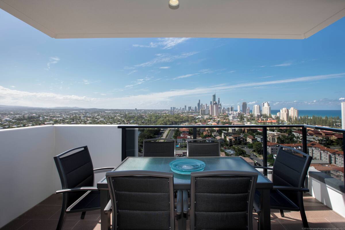 A balcony with a table and chairs, overlooking a cityscape with tall buildings and a view of the sea.
