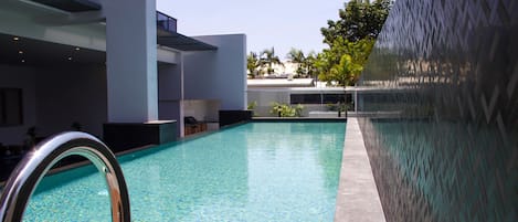 A modern indoor swimming pool with clear water, surrounded by a white building with balconies and a glass wall reflecting the sky.