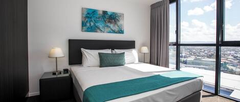 Una habitación de hotel con una cama grande, dos mesitas de noche, un ventilador de techo, vistas a la ciudad y un cuadro en la pared.