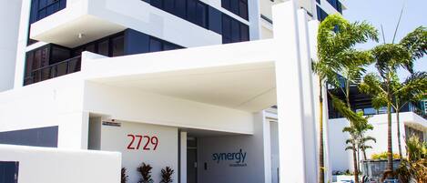 Um edifício moderno com o número 2729, uma placa com 'synergy broadbeach' e uma área ajardinada com palmeiras.