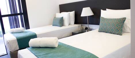 Habitación de hotel con dos camas, cada una con ropa blanca y cubrecamas color aguamarina. Entre las camas hay una mesita de noche con una lámpara.