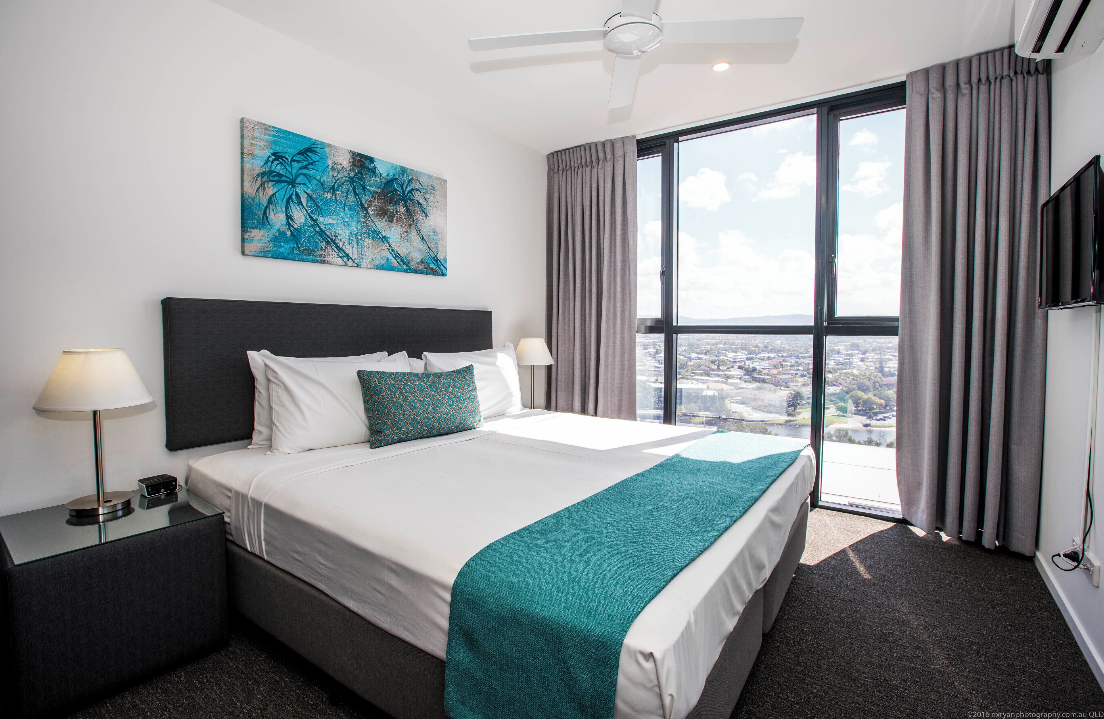 A hotel room with a large bed, a city view, a ceiling fan, and a flat-screen TV.