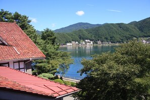 A lakeside view with a building featuring a red roof, surrounded by lush greenery and mountains in the background. - Kawaguchiko Hotel (Fujikawaguchiko)