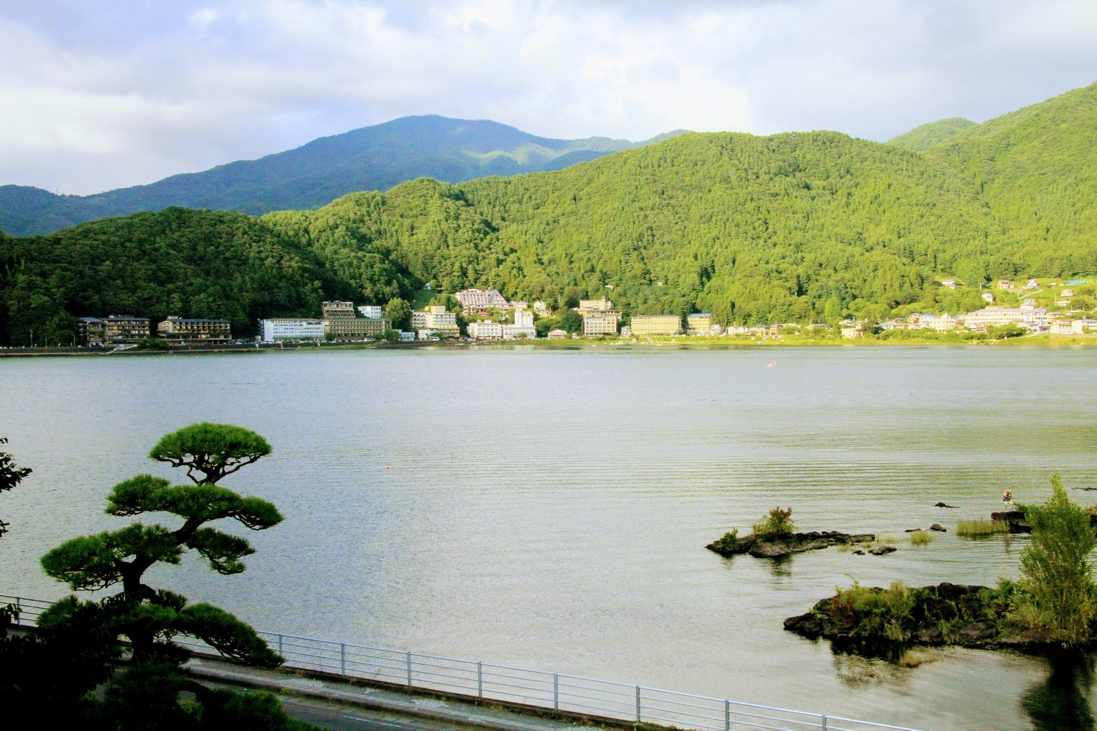 Un lago sereno con montañas al fondo, algunos edificios a la orilla y un pino destacado en primer plano.