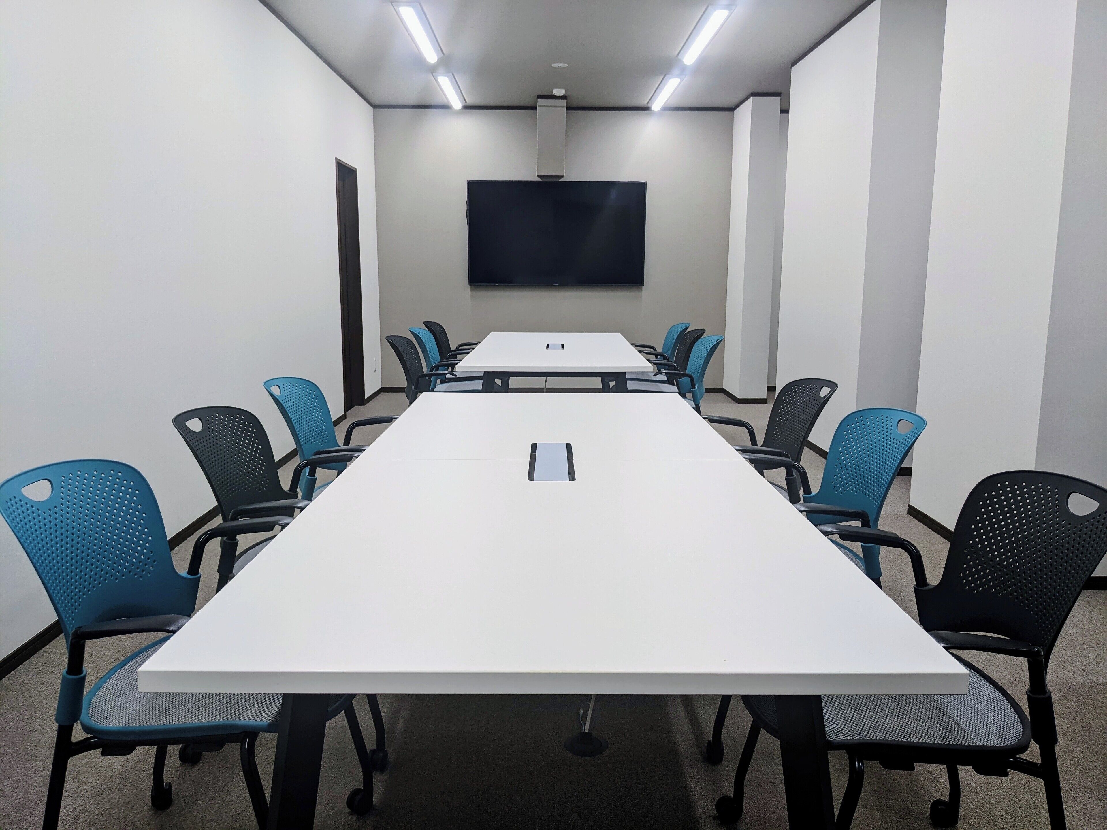 A conference room with a long table, blue and black chairs, a flat-screen TV on the wall, and a carpeted floor.
