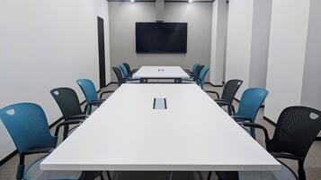 A conference room with a long table, blue and black chairs, a flat-screen TV on the wall, and a carpeted floor.