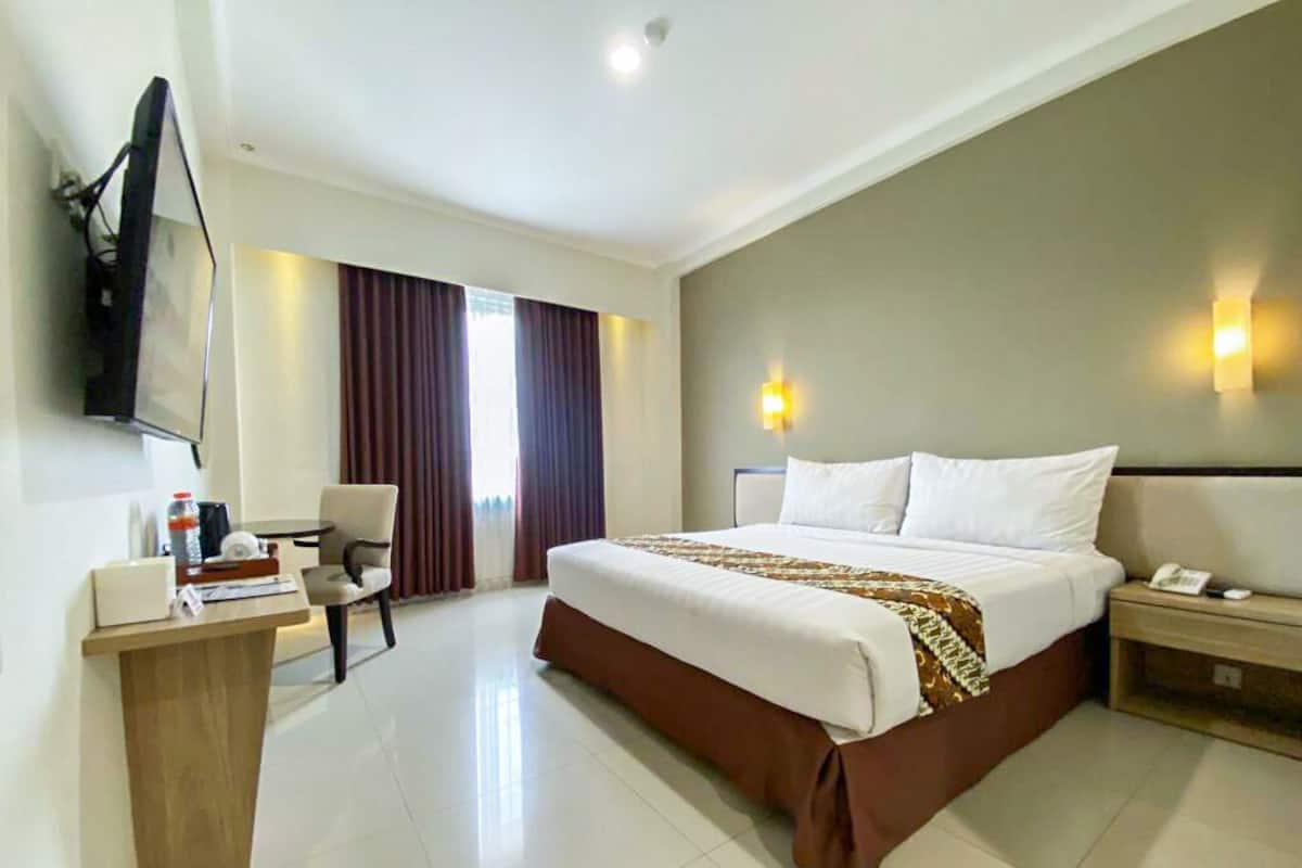Una habitación de hotel con una cama grande, un escritorio con computadora, una silla, un televisor montado en la pared y una ventana con cortinas.