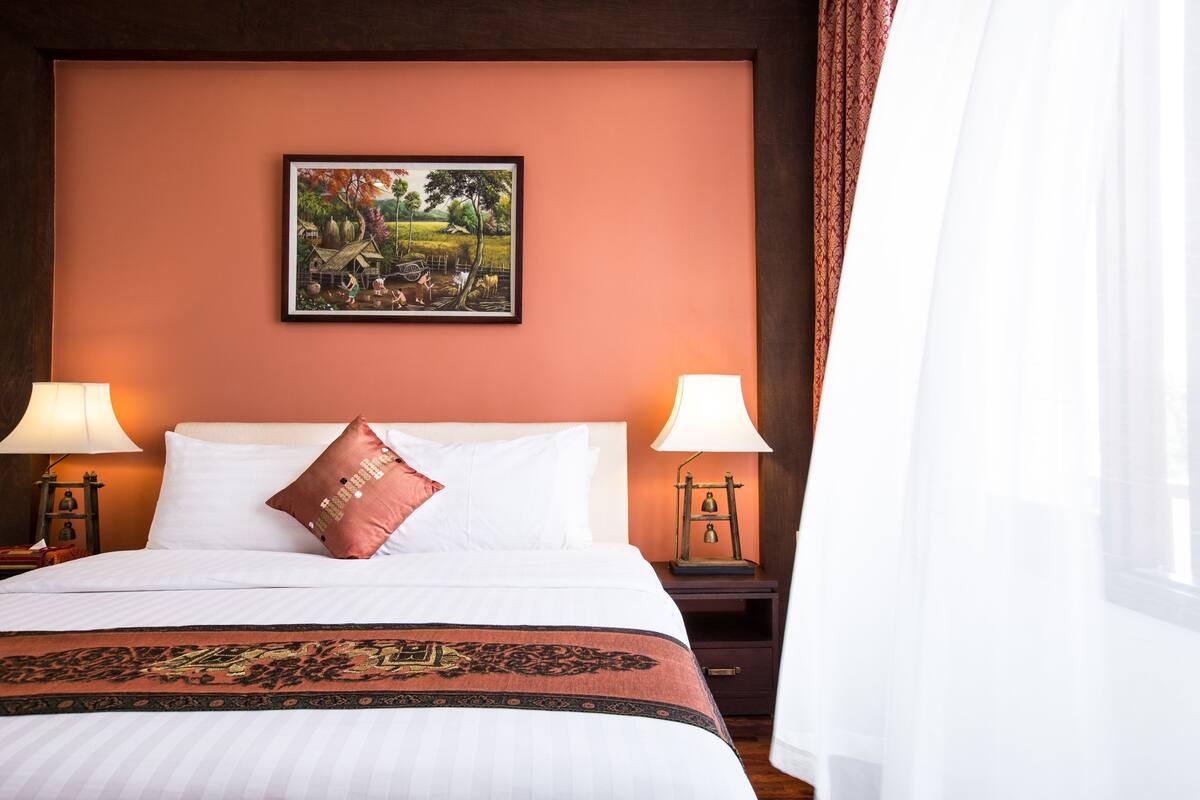 Habitación de hotel con una cama grande, dos lámparas de noche, un cuadro en la pared y una ventana con cortinas.
