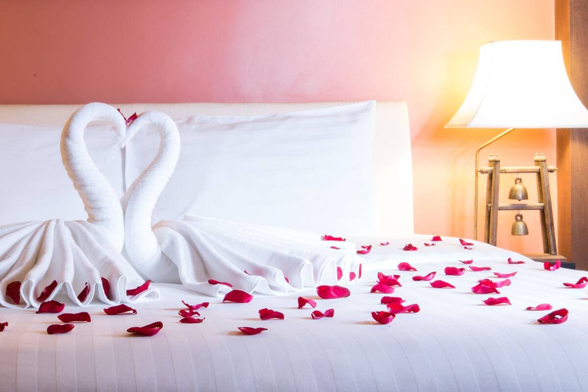Una cama con un cubrelecho blanco, dos lámparas fijadas a la pared y un cisne decorativo hecho con toallas en la cabecera.