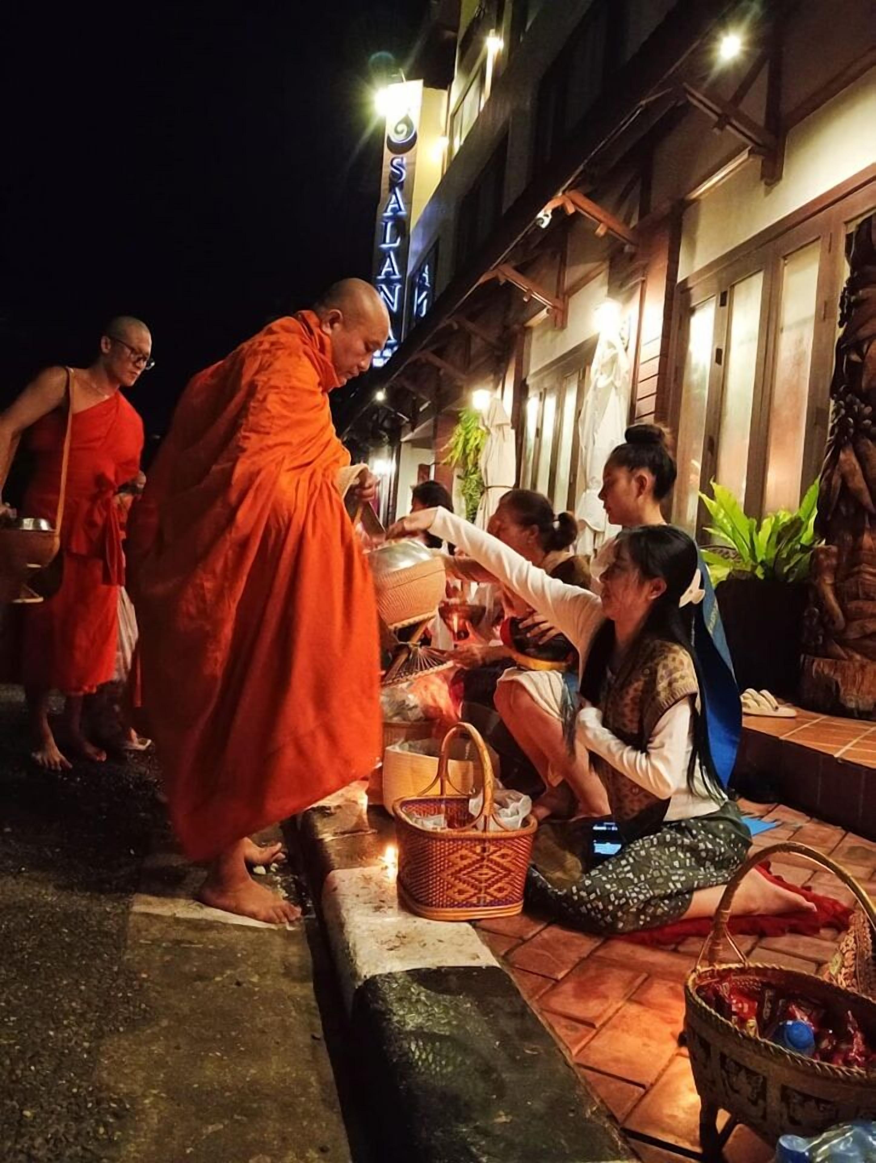 Un moine vêtu d’une robe orange échange avec des personnes devant un bâtiment sur lequel on peut lire l’inscription « SALAM ».