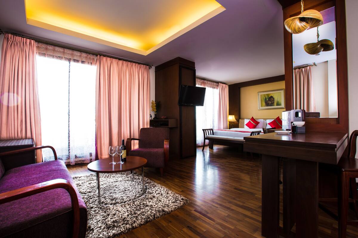 Habitación de hotel con sofá, una pequeña mesa con una botella de vino, una silla, cama, escritorio y televisión.