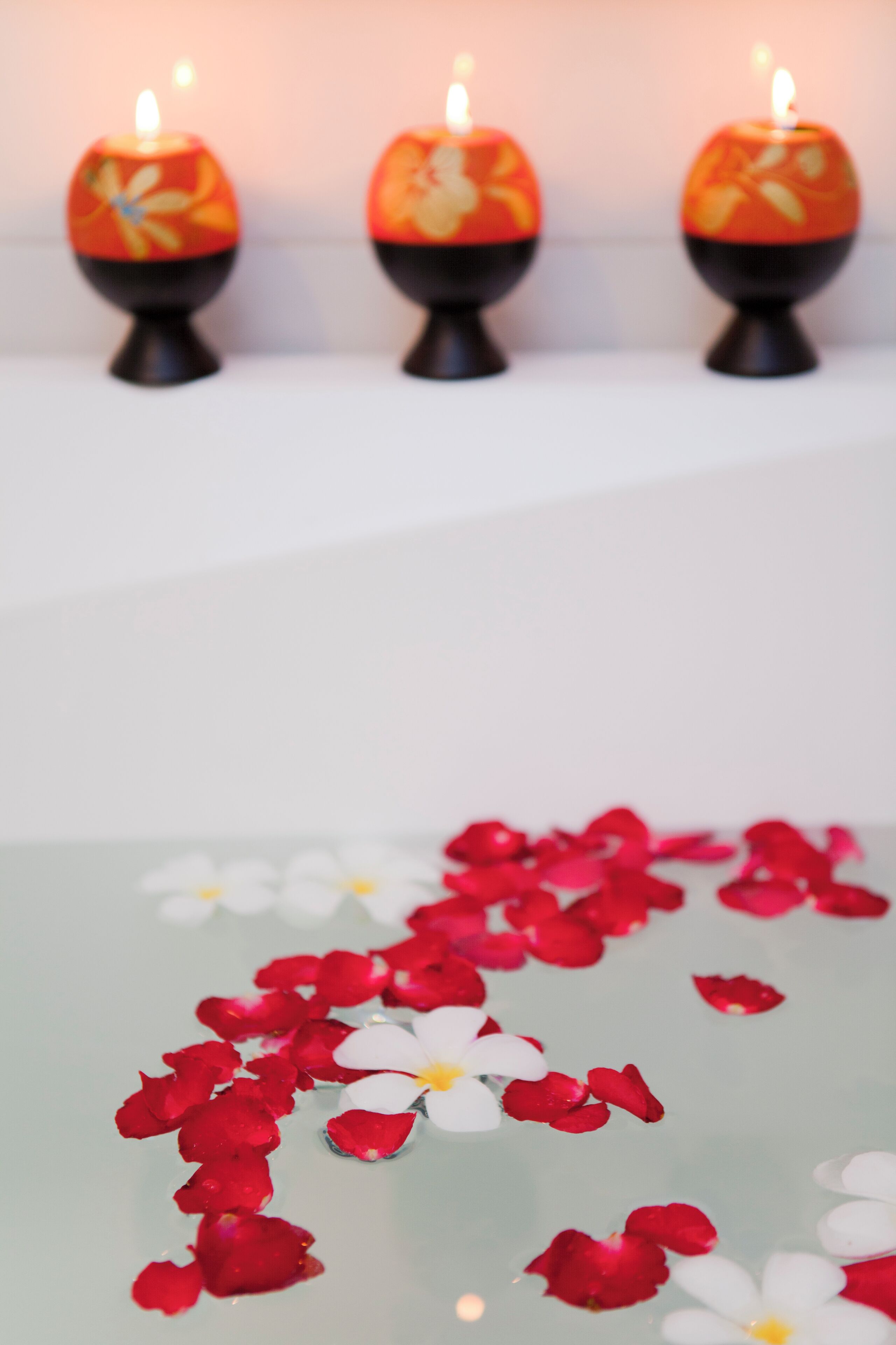 Three lit candles on a shelf, with rose petals scattered on a reflective surface below.