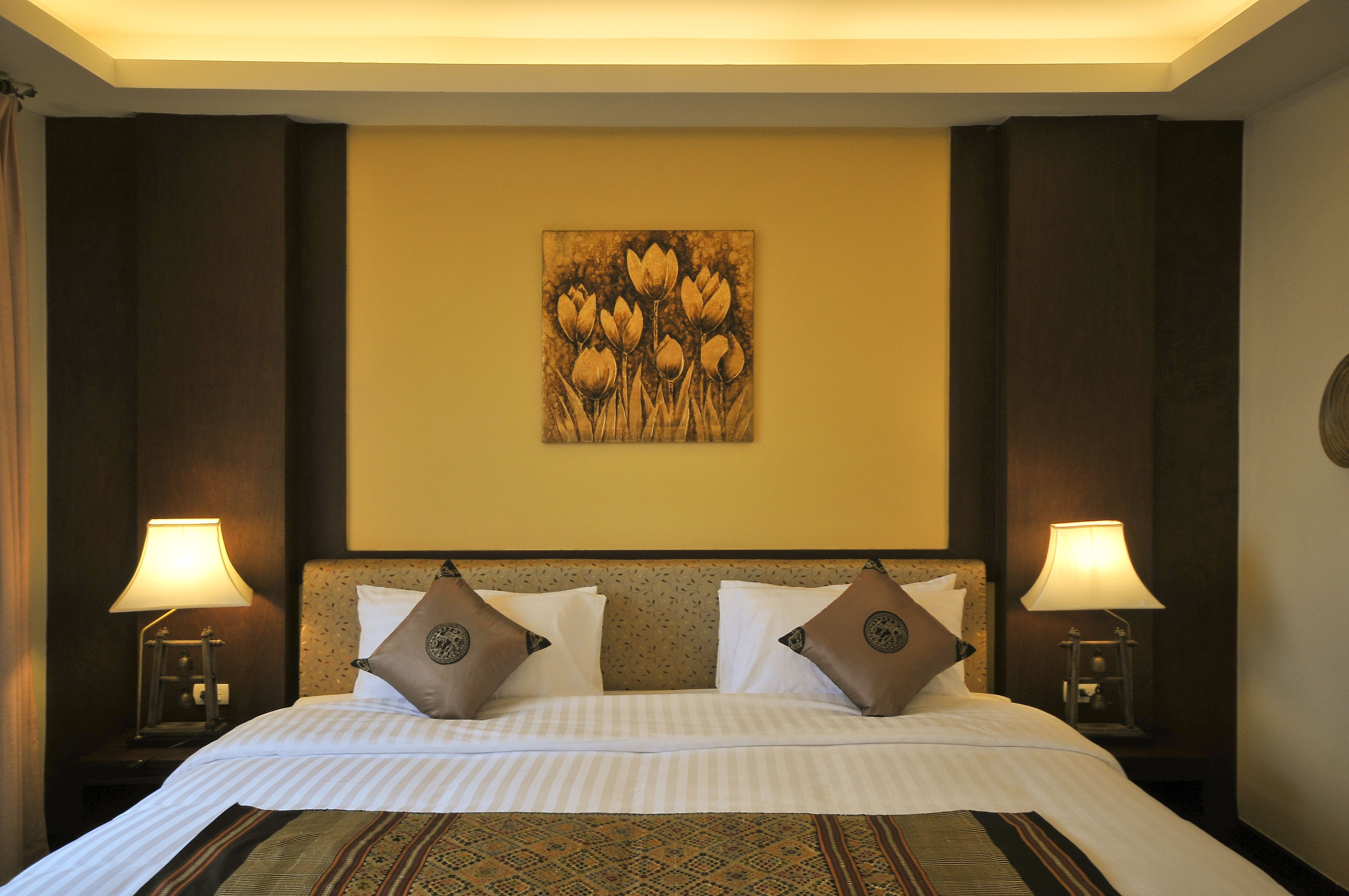A hotel room with a large bed, two bedside lamps, a headboard with a floral pattern, and a wall-mounted artwork of flowers.