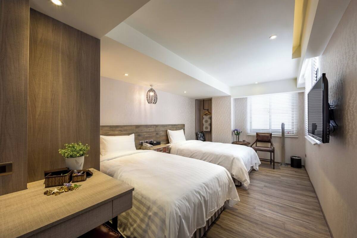 A modern hotel room with two beds, a wooden desk, a small table, a potted plant, and a television mounted on the wall.