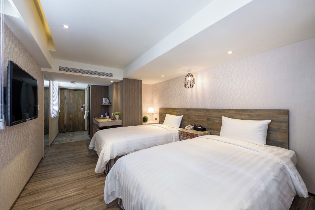 A hotel room with two beds, a TV, a bedside table with a lamp, and a wooden headboard.