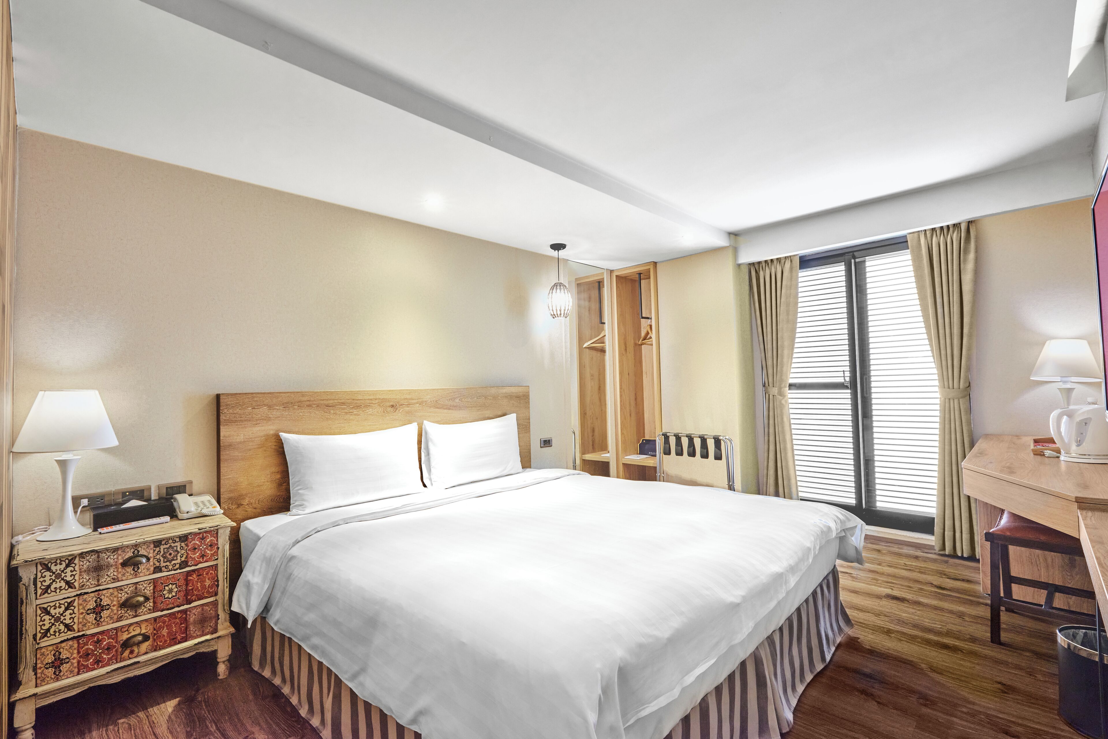 A hotel room with a large bed, a wooden nightstand, a desk, and a window with curtains.