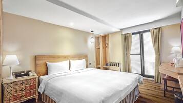 A hotel room with a large bed, a wooden nightstand, a desk, and a window with curtains.