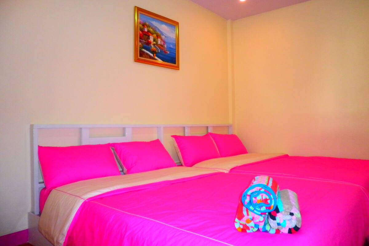 A bedroom with a large bed, white headboard, and bright pink bedding. There is a painting on the wall above the bed.