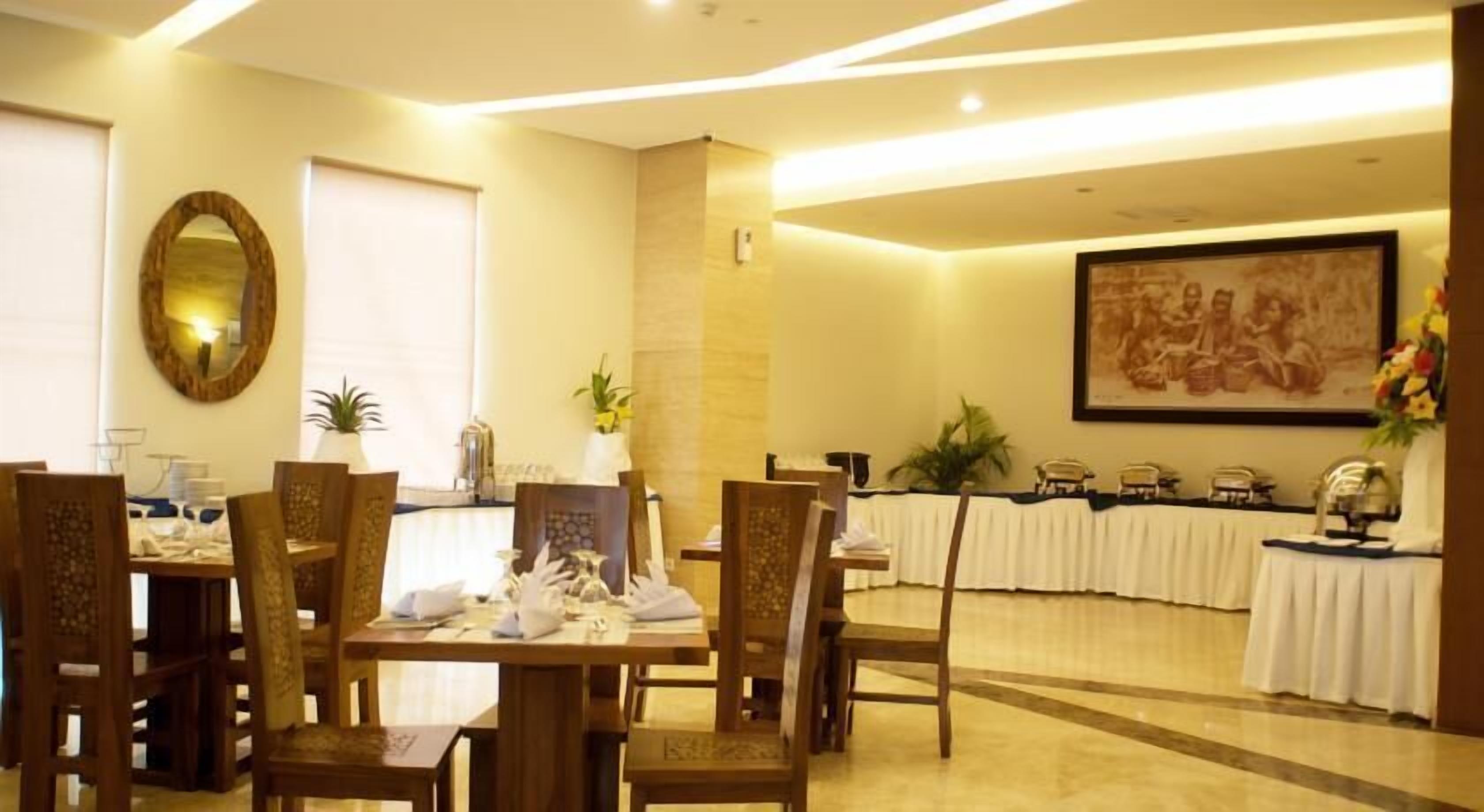 A hotel dining area with wooden chairs and tables, a large framed painting on the wall, and a buffet setup.
