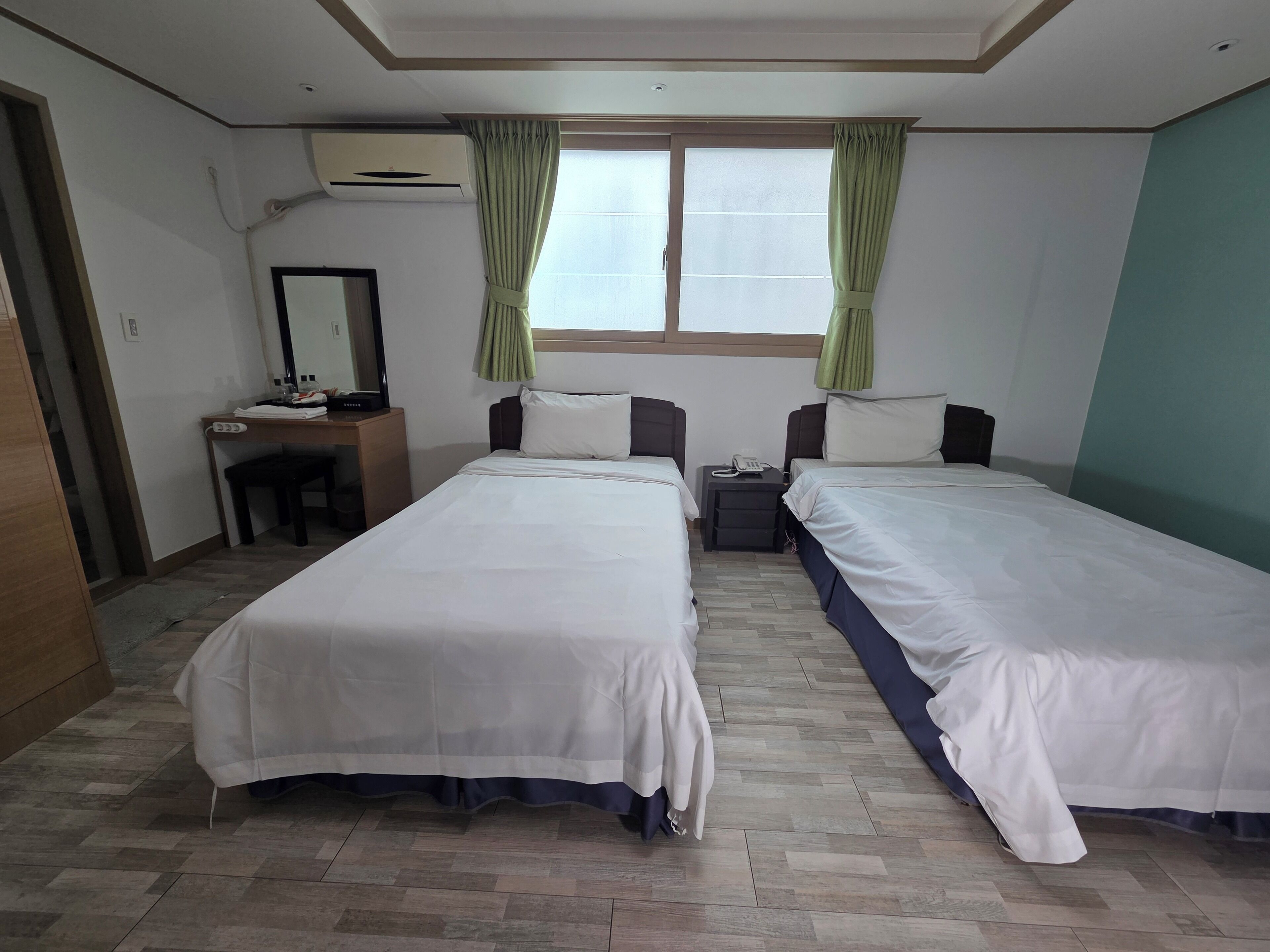 Una camera d'albergo con due letti singoli, una scrivania con una sedia, uno specchio e una finestra con tende.