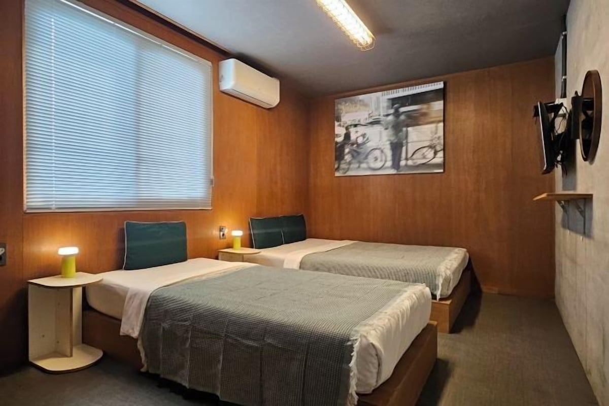 Habitación compacta con dos camas, una ventana con persianas, aire acondicionado montado en la pared, una mesita de noche pequeña con lámpara y un cuadro enmarcado en la pared.