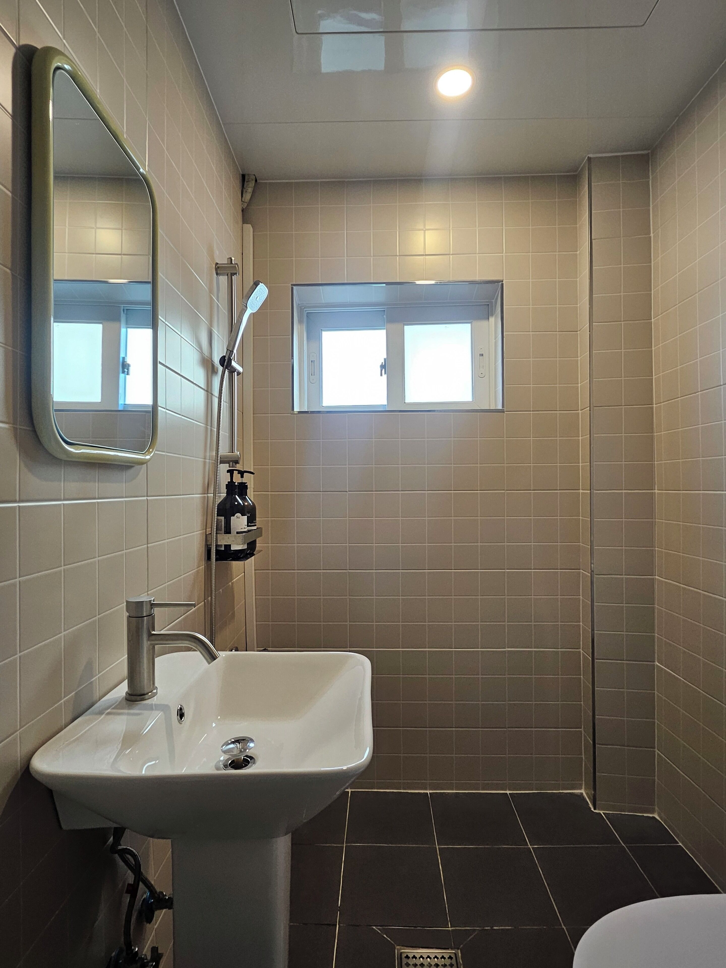 A bathroom with a sink, mirror, shower, and a small window.