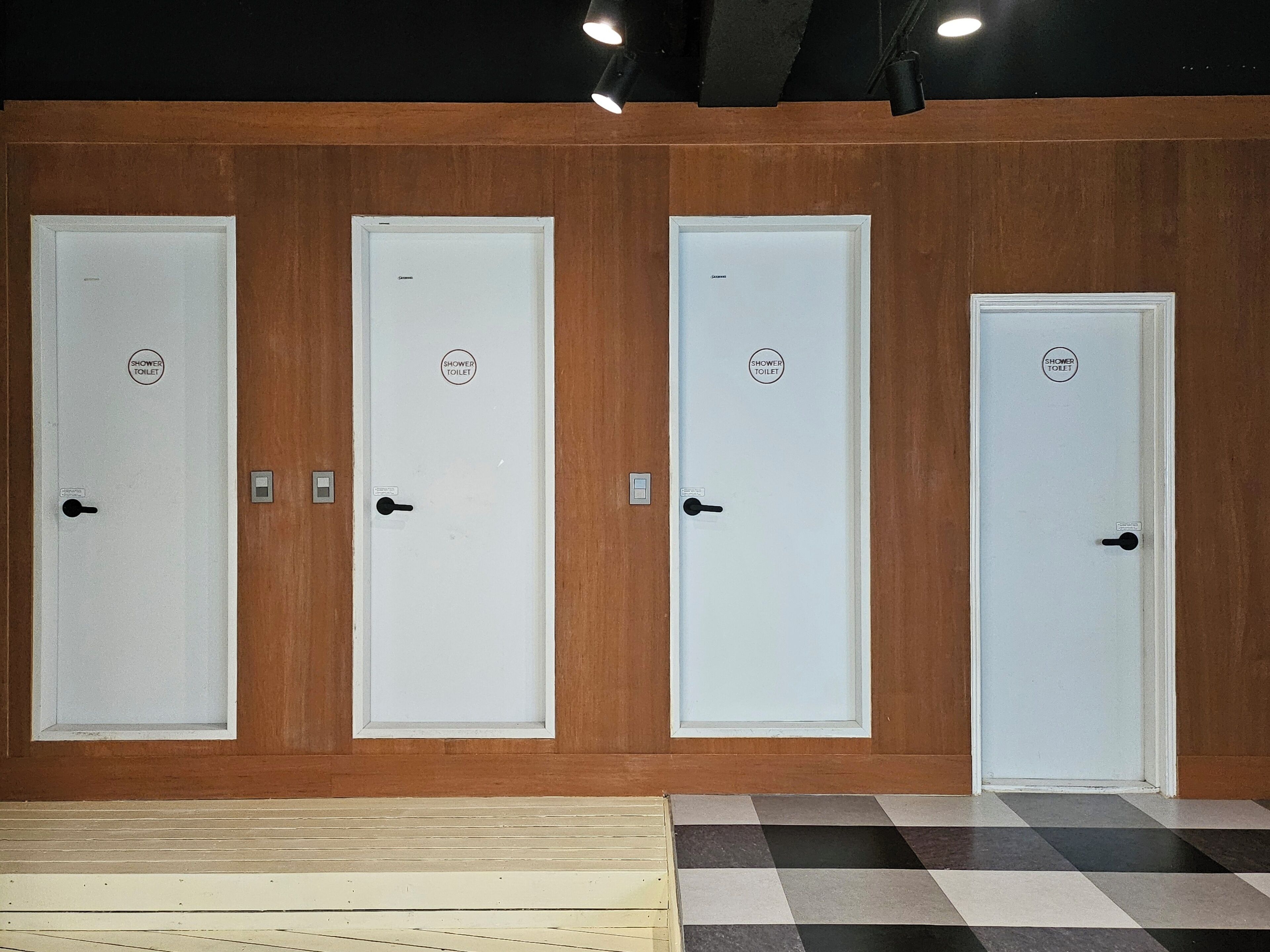 A row of four white doors with "SHOWER TOILET" labels, set against a wooden wall with a checkered floor.