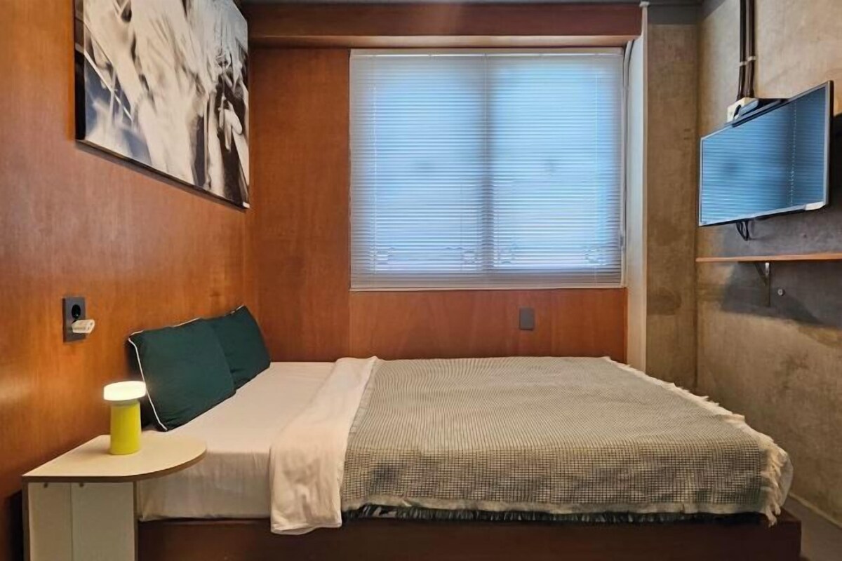 Una habitación de hotel con cama, mesita de noche con lámpara, ventana con persianas y televisor montado en la pared.