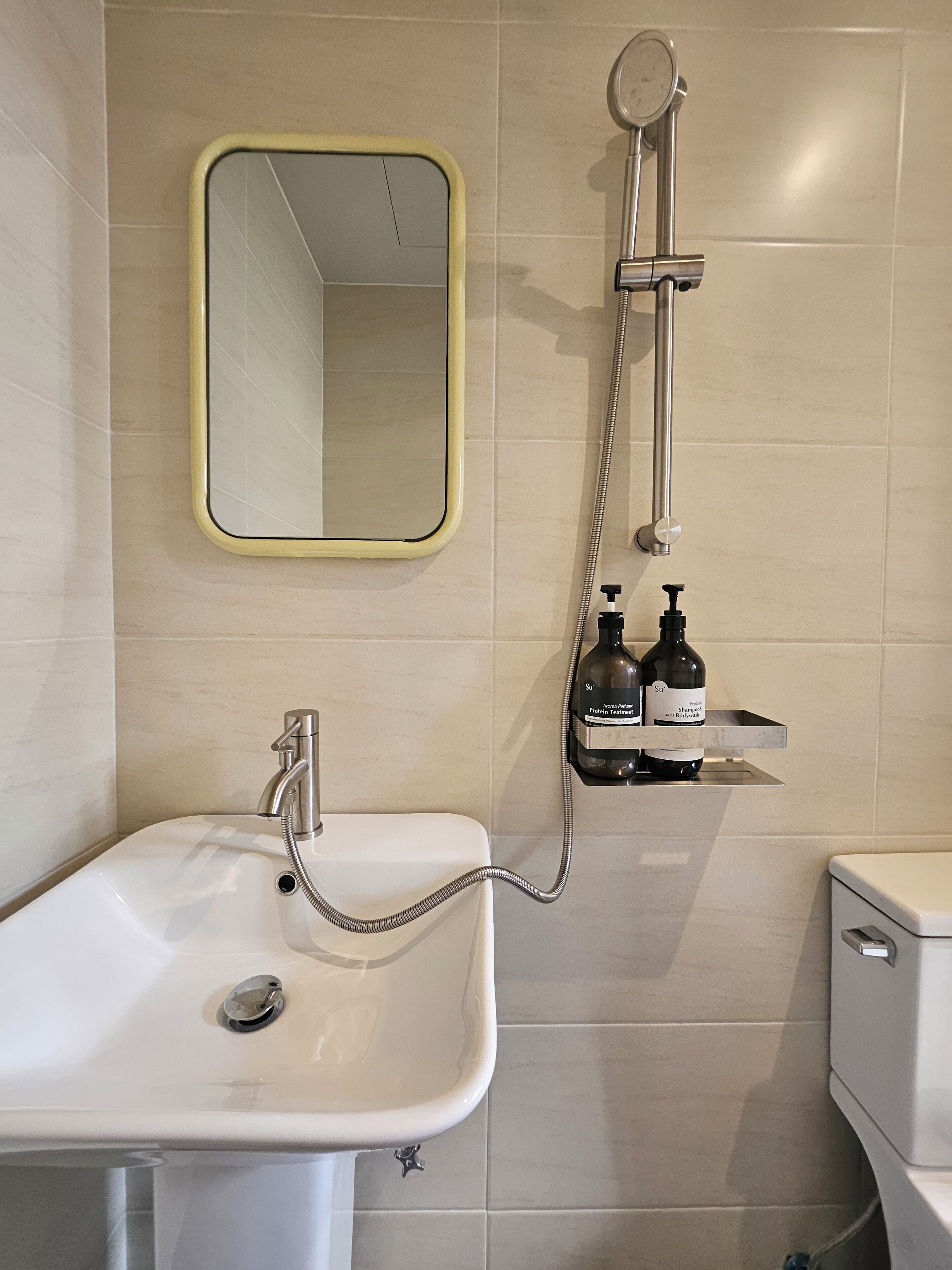 A bathroom with a white sink, a mirror, a shower, and two bottles of shampoo on a shelf.
