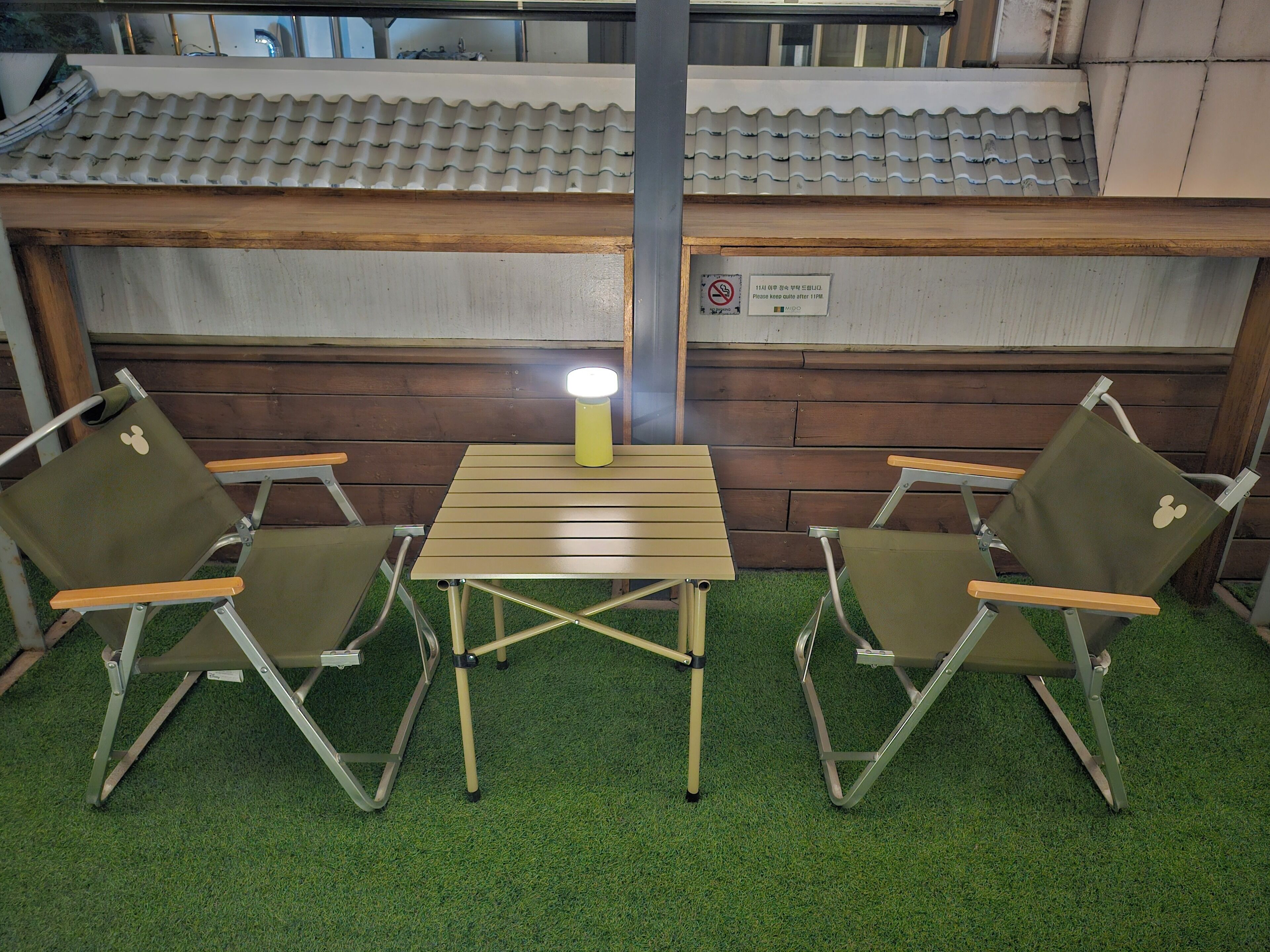 Un petit espace extérieur aménagé avec deux chaises pliantes et une table, une lampe et un panneau fixé au mur.