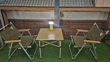 Un petit espace extérieur aménagé avec deux chaises pliantes et une table, une lampe et un panneau fixé au mur.